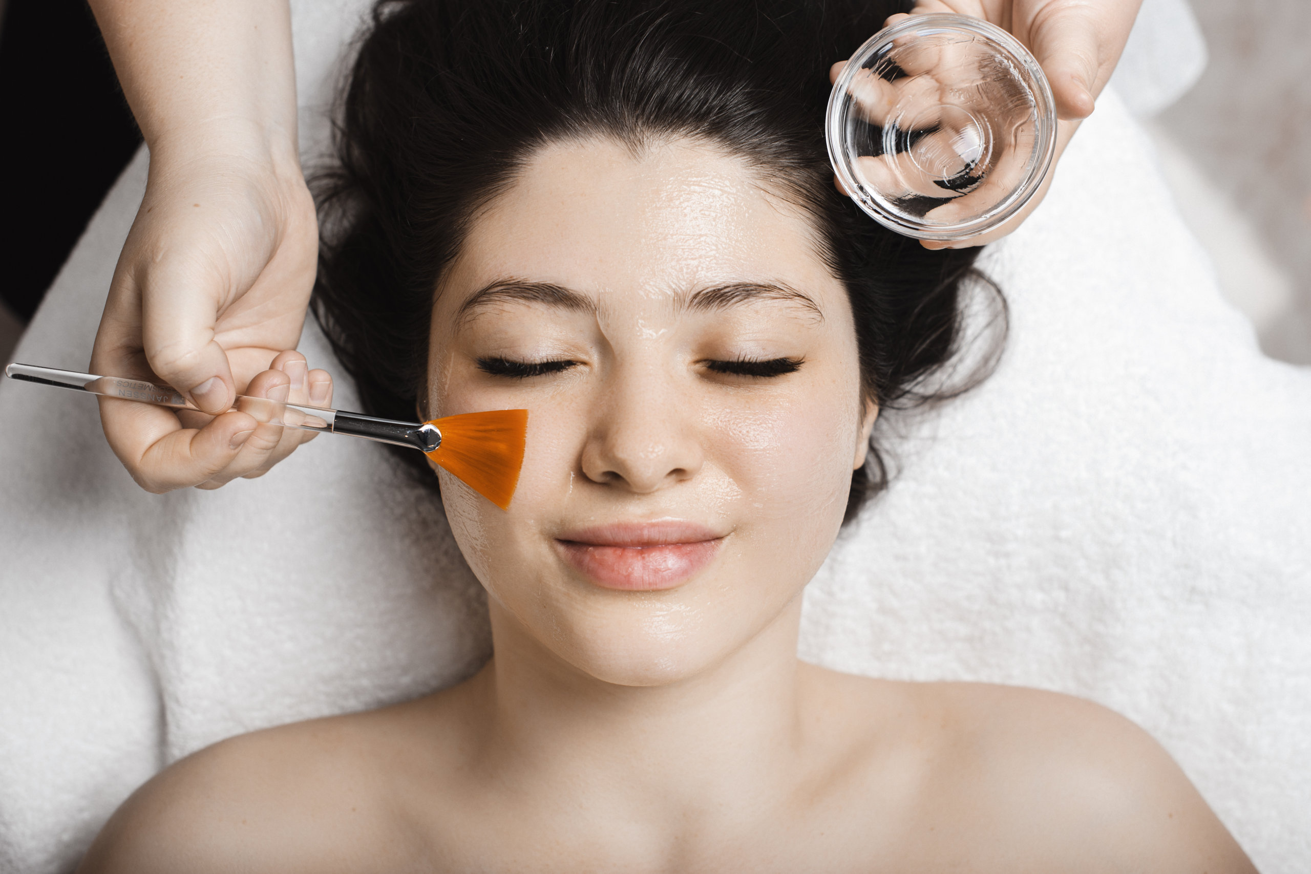 Obere Ansicht einer schönen Frau, mit geschlossenen Augen, während eine Kosmetikerin die Stammzellenmaske auf ihr Gesicht aufträgt